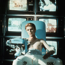 David Bowie sitting in a chair in front of TV screens