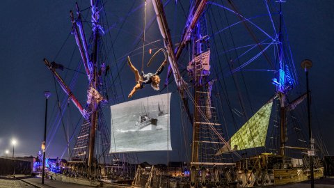 Celluloid Sail - tall ship docked to harbourside with projections on sails and acrobat hanging from rope.