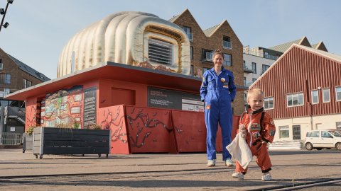 A lady in a blue jumpsuit stands with her hands in her pockets before a large gold domed building - a martian house. A child in an orange jumpsuit stands in the foreground.
