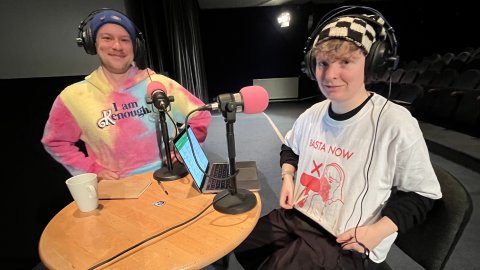 Stef and Steph sit at a table for podcast in Cinema One. They are showing off their respective tops to the camera. Stef is wearing an 'I am Kenough' hoodie. Steph is wearing a Basta now t-shirt.