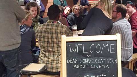 Deaf Conversations About Cinema People engaged in conversation around a table in the cafe bar in front of a sign reading Welcome to Deaf Conversations About Cinema