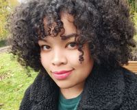 Malaika is a mixed race (Black African & White) cis woman. She has shoulder length dark, curly, afro hair with a fringe. She is wearing a green jumper dress with a fluffy black coat over the top. She smiles at the camera, with light red lipstick on.