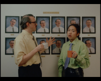 a still from a music video. Two colleagues appear to be stood in front of an employee of the month picture frame wall. The pictures are all of the same man