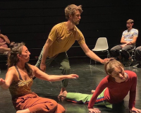 A colour photo of three performers  Kip,  Linzy  and Holly in a black box theatre space.  The are wearing orange yellow and green clothing, Kip is standing.  Linzy and Holly are close to the ground. The trio in mid movement use arms legs and undulating spines like a salamander moving through water, sensing their surrounding through the surface of their skin. 