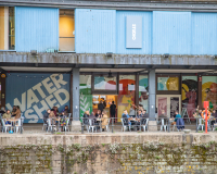 A photo of the external area of Watershed, focusing on the Undershed. Large illustrated artwork by Lucy Turner is on the windows and people sit around tables outside on the Harbourside