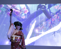 A woman dressed in traditional Japanese clothing, in a VR headset, plays with a VR performance piece, projected onto a screen behind her. 