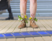 A photo of a pair of legs from the knee’s down, with the feet adorned in brown boots brimming with flowers. In the background you can see the silver shutter of a shop and the legs of another person walking past.