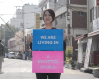 Image of Sooah Kwak standing in the street in South Korea holding a sign that says We Are Living In A Knotted World