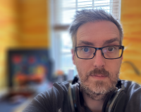 A middle age man with greying hair, glasses, a beard and headphones around his neck