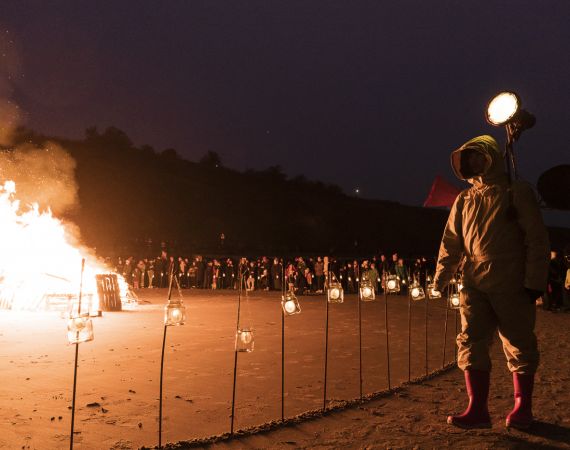 Photo from Beacons project in Folkestone. A figure stands on a beach after dark with flames and candles behind them. An audience can be seen gathered in the background