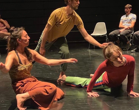 A colour photo of three performers  Kip,  Linzy  and Holly in a black box theatre space.  The are wearing orange yellow and green clothing, Kip is standing.  Linzy and Holly are close to the ground. The trio in mid movement use arms legs and undulating spines like a salamander moving through water, sensing their surrounding through the surface of their skin. 