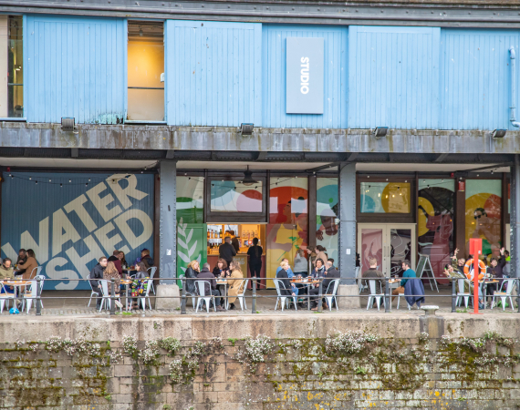 A photo of the external area of Watershed, focusing on the Undershed. Large illustrated artwork by Lucy Turner is on the windows and people sit around tables outside on the Harbourside