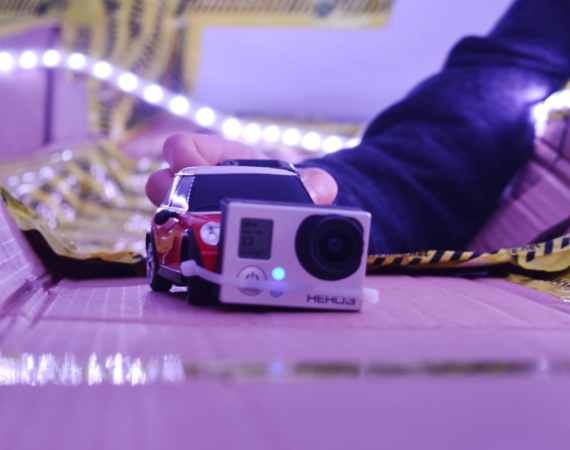 A child's hand places a remote control car with a go pro camera attached to the front of it onto a self built cardboard racetrack held together by tape with led strip lights attached in a DIY manner