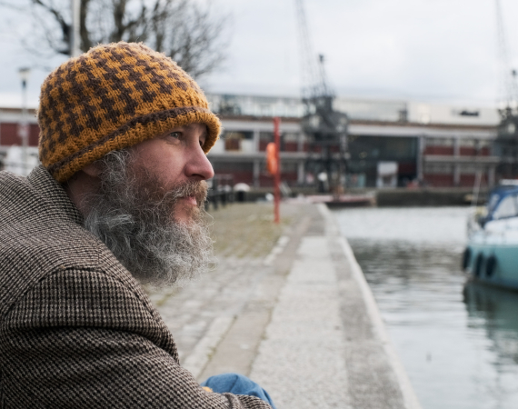 A white man with a beard and a yellow wool hat looks out over the harbourside in Bristol