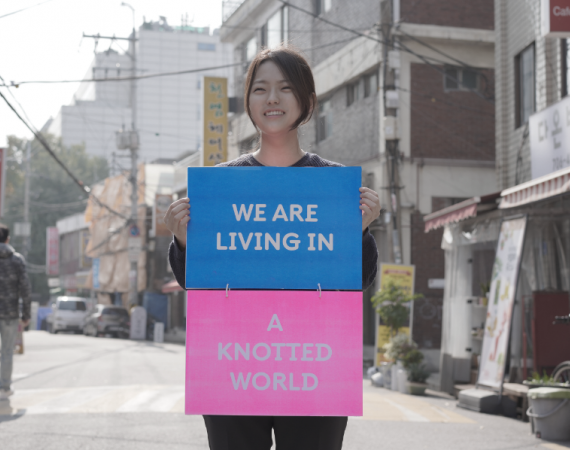 Image of Sooah Kwak standing in the street in South Korea holding a sign that says We Are Living In A Knotted World
