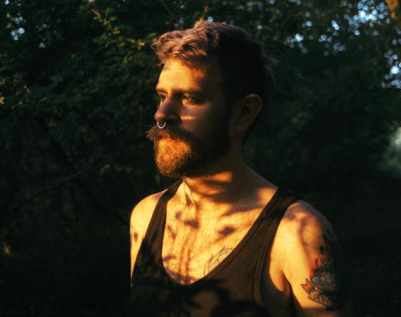 Graham is a person with a beard and a mohawk. He is stood in a dimly lit forest wearing a black vest.