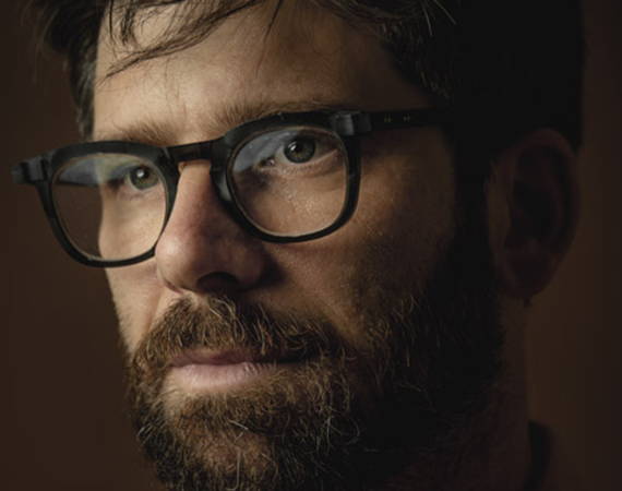 Portrait of Ben Samuels, a person with a beard and glasses, set against a dark backdrop.