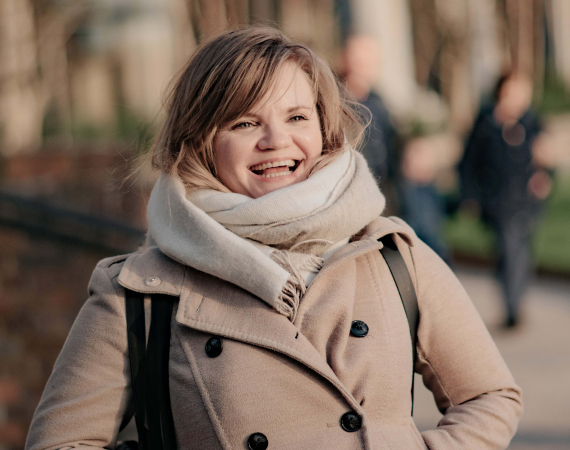 A blonde woman in her 30s laughs in a beige winter coat and cream scarf.