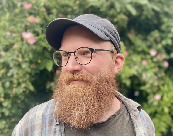 Headshot of me in front of green shrubs. I have a large ginger beard, and am wearing glasses, a black baseball cap, and a faded checked shirt