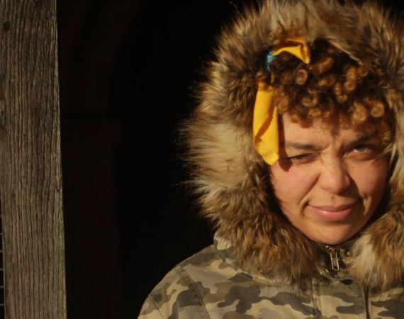 A women of colour squinting into the sun. She is wearing a green camouflage coat, with a fur trim around her face and her yellow hair tie  is visible through her curly afro.
