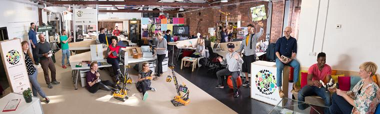 A number of people spread around Pervasive Media Studio, each doing different things such as chatting with colleagues, showing work to colleagues, or posing with some of their work.