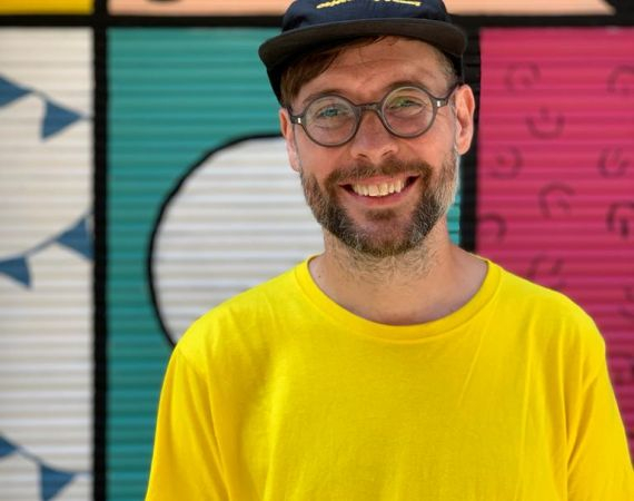 Anders, a light-skinnned bearded person, stands infront of a colourful background. We can see his head and shoulders. He is smiling and wearing a black cap, glasses and a bright yellow t-shirt. 