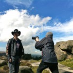 K+Jex Colborne, in a leather jacket and stetson, is being filmed by a camera person against a dramatic backdrop of Yorkshire moors and cloud filled blue sky