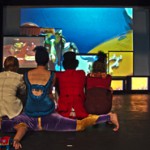 box_of_frogs_all_on_kaveh-copy the cast of Box of Frogs are sitting, backs to the audience, upon one of the performers - they are all watching a big screen upon which we can see circus Playmobile