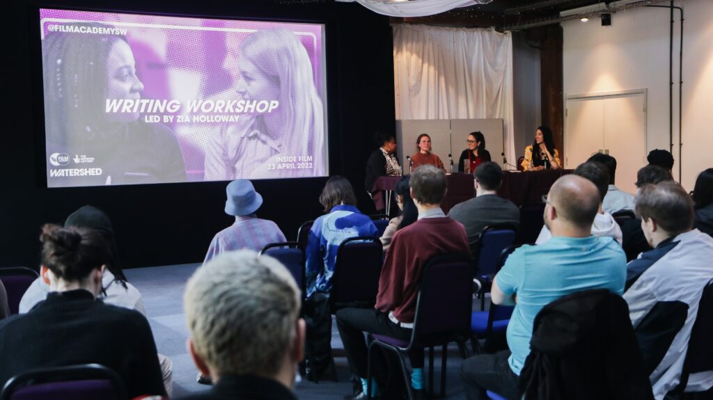 A room of seated people. At the front a panel of four women are seated at a table with microphones and behind them is a purple slide that has 'writing workshop' written on them