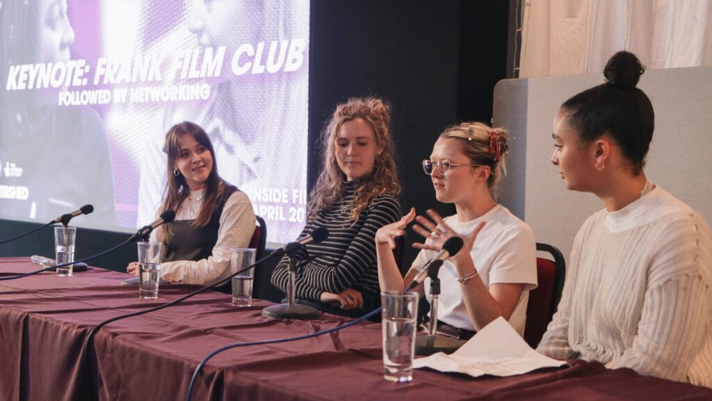 A panel of four women sat in a row at a table, one of whom is speaking. Behind them is a purple slide that reads "Kenyote: Frank Film Club"