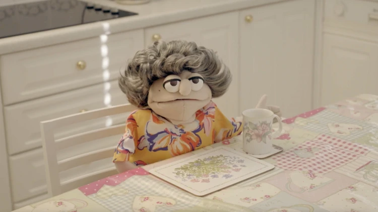 Image: Gender Bender dir. Grace Groves A puppet with grey curly hair, floral shirt, sat at a table