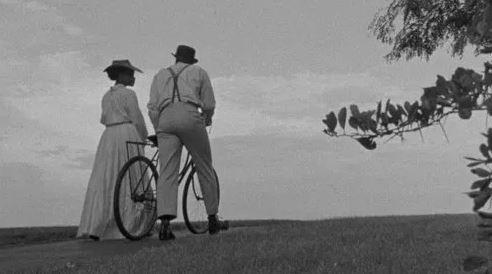 Black-and-white still from Compensation. The back of two people walking in the park next to a tree, one pushing a bicycle.