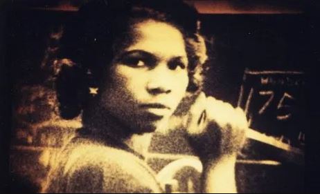 Black-and-white still from Handsworth Songs, a person looking at the camera stood in front of a blackboard, gripping an item