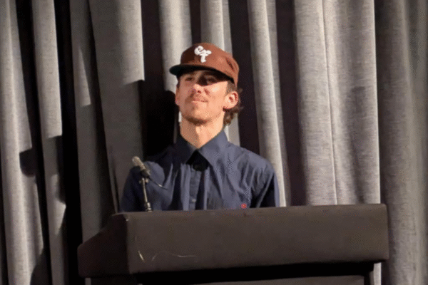 Host Jojo Myers-Insole stood at a lecturn wearing a brown cap and dark blue hat
