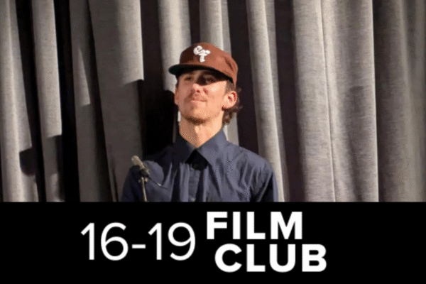 Host Jojo Myers-Insole stood at a lecturn wearing a brown cap and dark blue hat. 16-19 Film Club logo