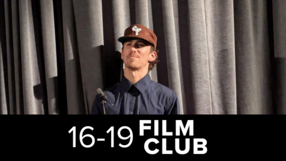 Host Jojo Myers-Insole stood at a lecturn wearing a brown cap and dark blue hat. 16-19 Film Club logo