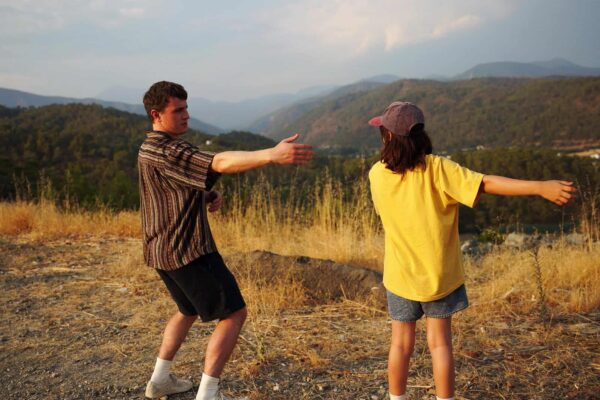 Still from Aftersun of a man wearing a striped shirt and shorts with a girl wearing a yellow t-shirt and shorts perform tai-chi on a grassy hill