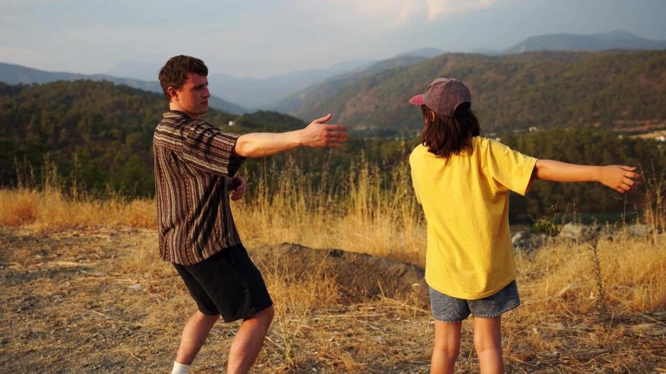 Still from Aftersun of a man wearing a striped shirt and shorts with a girl wearing a yellow t-shirt and shorts perform tai-chi on a grassy hill