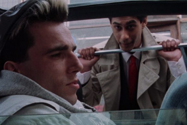 Daniel Day-Lewis and Gordon Warnecke in My Beautiful Laundrette. Gordon Warnecke is peering through a car window at Daniel Day-Lewis, sat down.