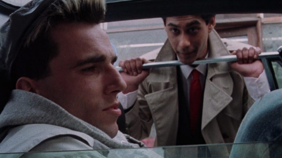 Daniel Day-Lewis and Gordon Warnecke in My Beautiful Laundrette. Gordon Warnecke is peering through a car window at Daniel Day-Lewis, sat down.