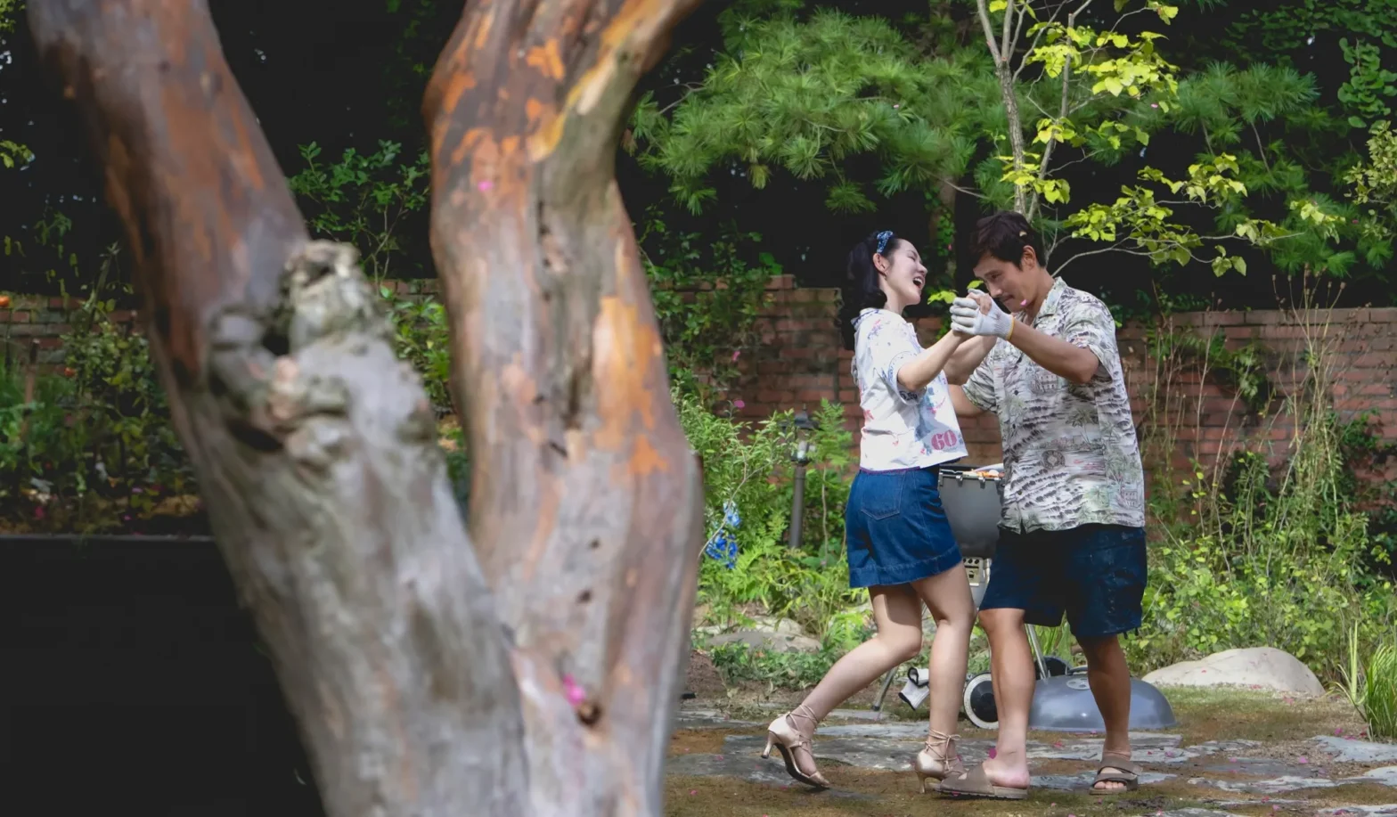 Still from No Other Choice of two people dancing in a park, with them in the background and a tree in the foreground of the shot.