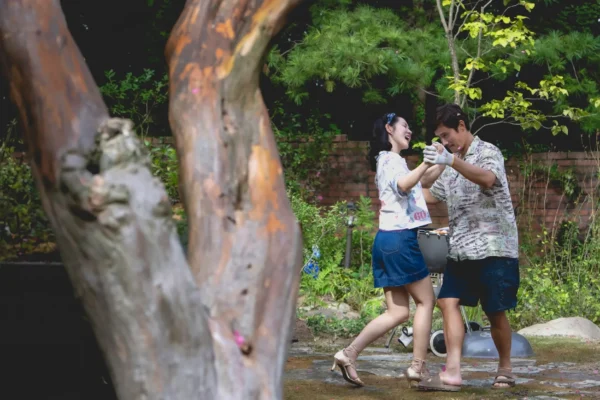 Still from No Other Choice of two people dancing in a park, with them in the background and a tree in the foreground of the shot.