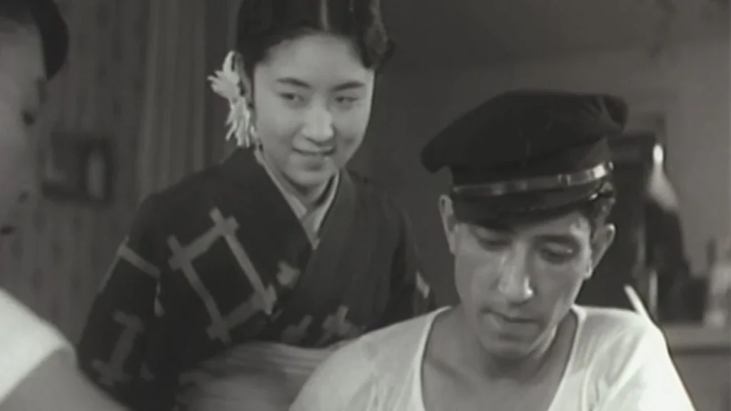 A woman smiles, watching, stood over a man wearing a hat looking down.