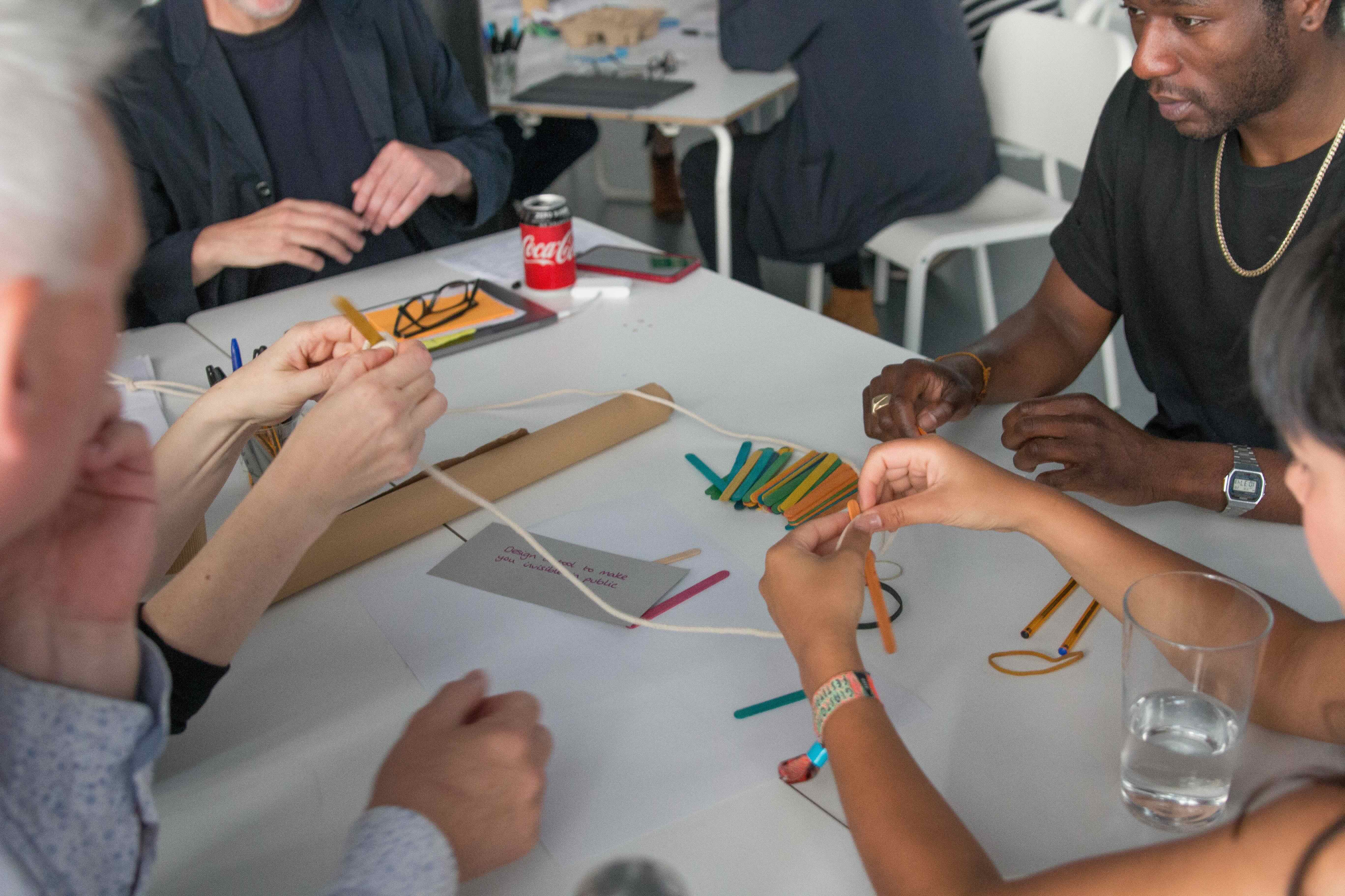 A diverse group if people are doing an activity together on a white tables with string and colourful lollipop sticks.