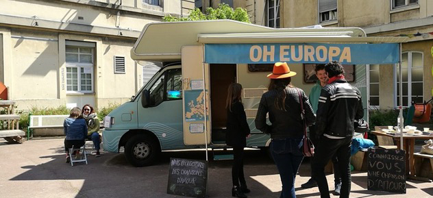 Old camper van in town square with some people looking at it.