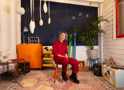 Image of Artist, Natalie Whitney in a red jumpsuit and black boots, they are sat smiling on a red stool.