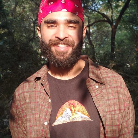 A young man of colour is wearing a red bandana and a brown shirt and t-shirt and smiles at the camera.