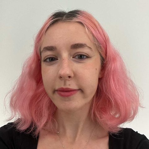 A white woman with wavy pink hair looks into the camera with a slight smile on her face.