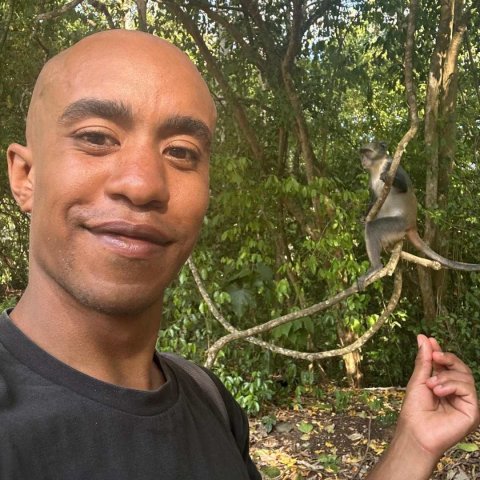 A young brown man is stood on the right with a slight smile, on the left he has his hand underneath a monkey.