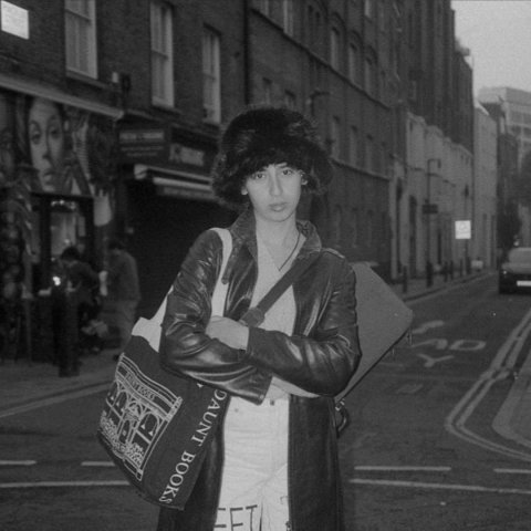 In the middle of an empty street a young woman looks into the camera wearing a leather jacket and a furry hat, the image is black and white.